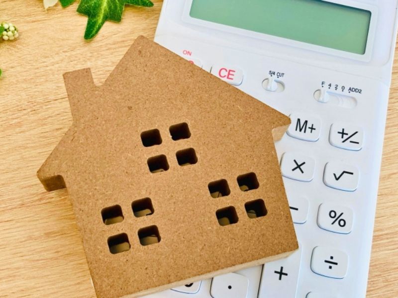 木製の家のオブジェと白い電卓が木目のテーブルの上に並んでいるイメージ画